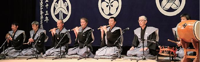 写真:下野の囃子方の皆さん。舞台右側から、2種類の太鼓、謡い、笛、鐘の順に、7人の男性が裃を身に着け座っています。