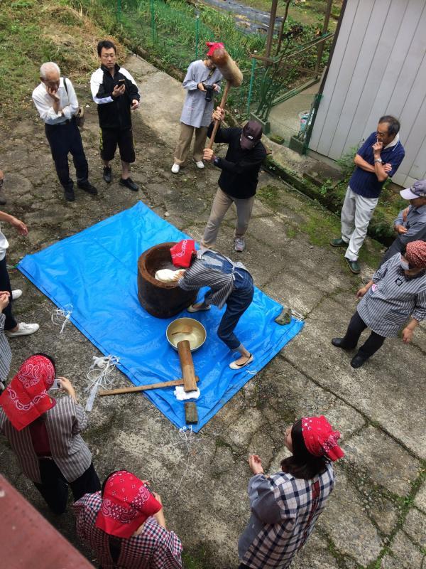 屋外でブルーシートを敷きその上で杵と臼で餅つきをする人たちの写真