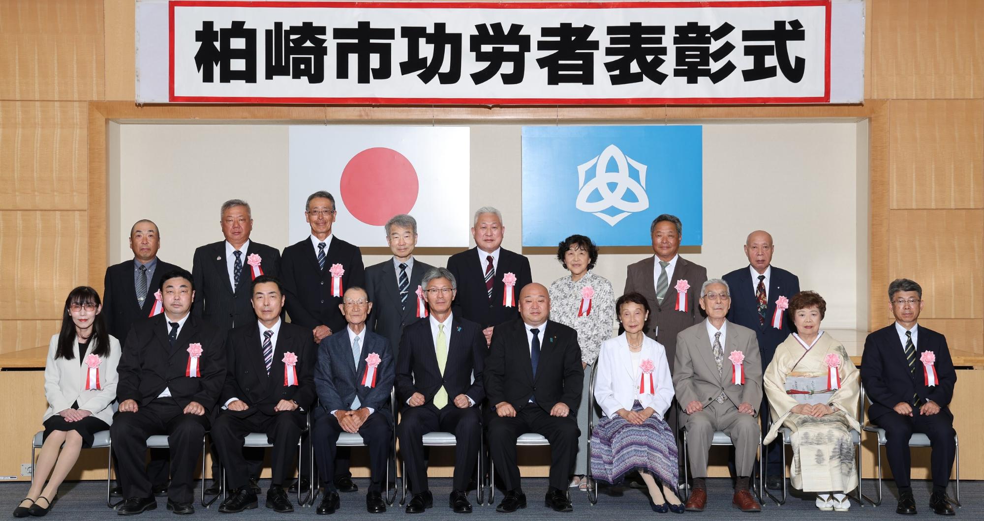 「柏崎市功労者表彰式」と書かれた横看板の下に国旗と市旗が掲げられ、その前に胸にリボンを付けた16人の受賞者と市長、議長が2列に並んで集合写真を撮っている