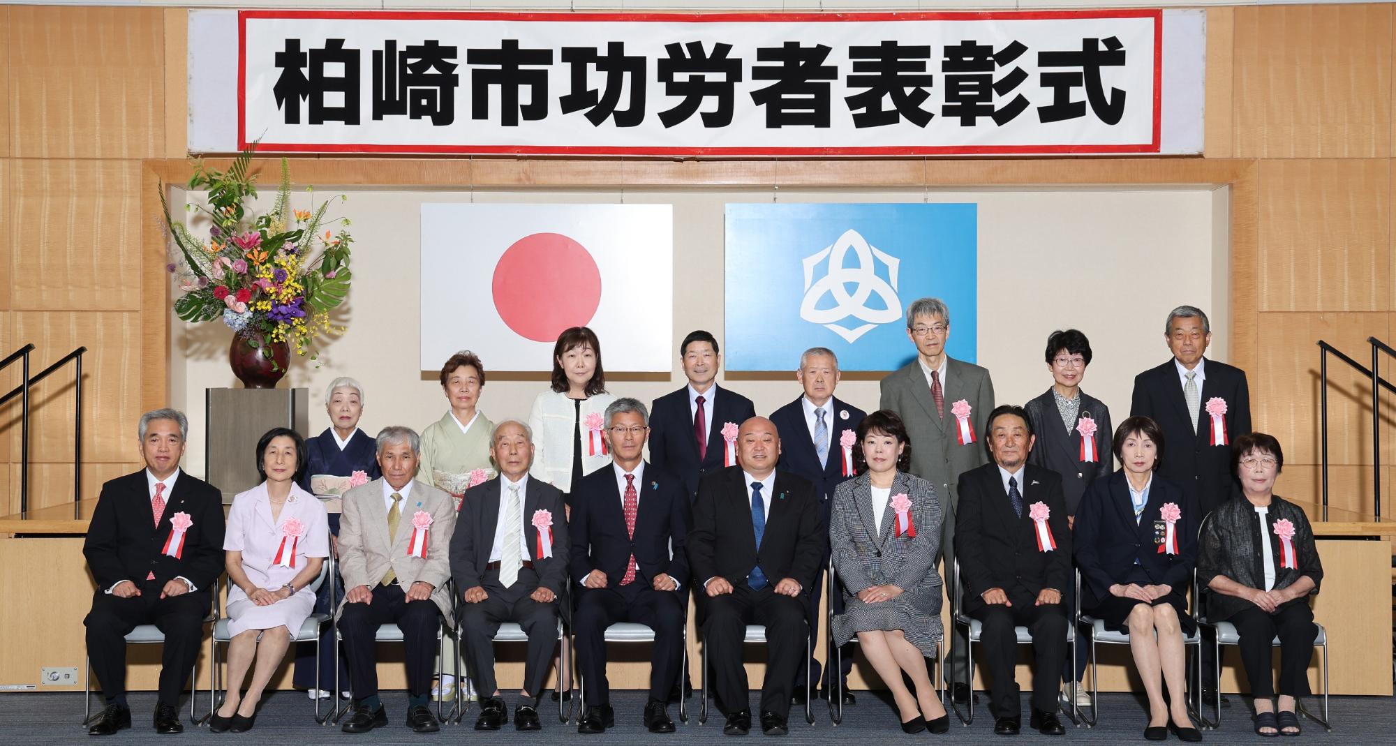 「柏崎市功労者表彰式」と書かれた横看板の下に国旗と市旗が掲げられ、その前に胸にリボンを付けた16人の受賞者と市長、議長が2列に並んで集合写真を撮っている