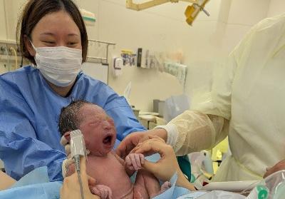 写真:青い病衣服を着た女性が、分娩直後の赤ちゃんを支え、産婦に見せているところ。赤ちゃんは大きな口を開けて泣いています