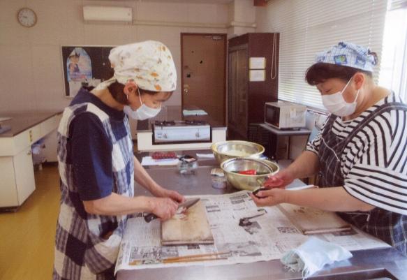 写真:調理室で2人の女性がナスの皮をむいたり、野菜を千切りしたりしています。