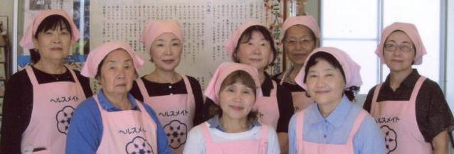 ピンク色のエプロンと三角巾をを身に着けた二中地区食生活改善推進員の女性8名中3名が前方に、後方に5名立っている写真