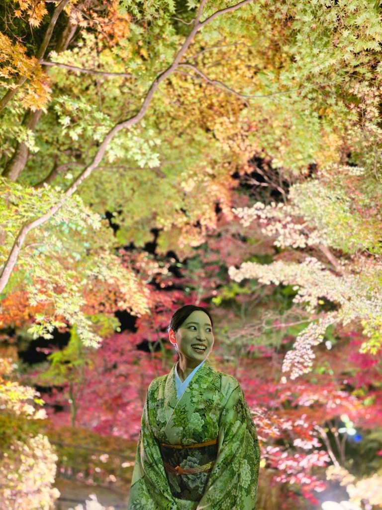 写真:女性がライトアップされた紅葉を背景に着物を着て笑顔を浮かべています