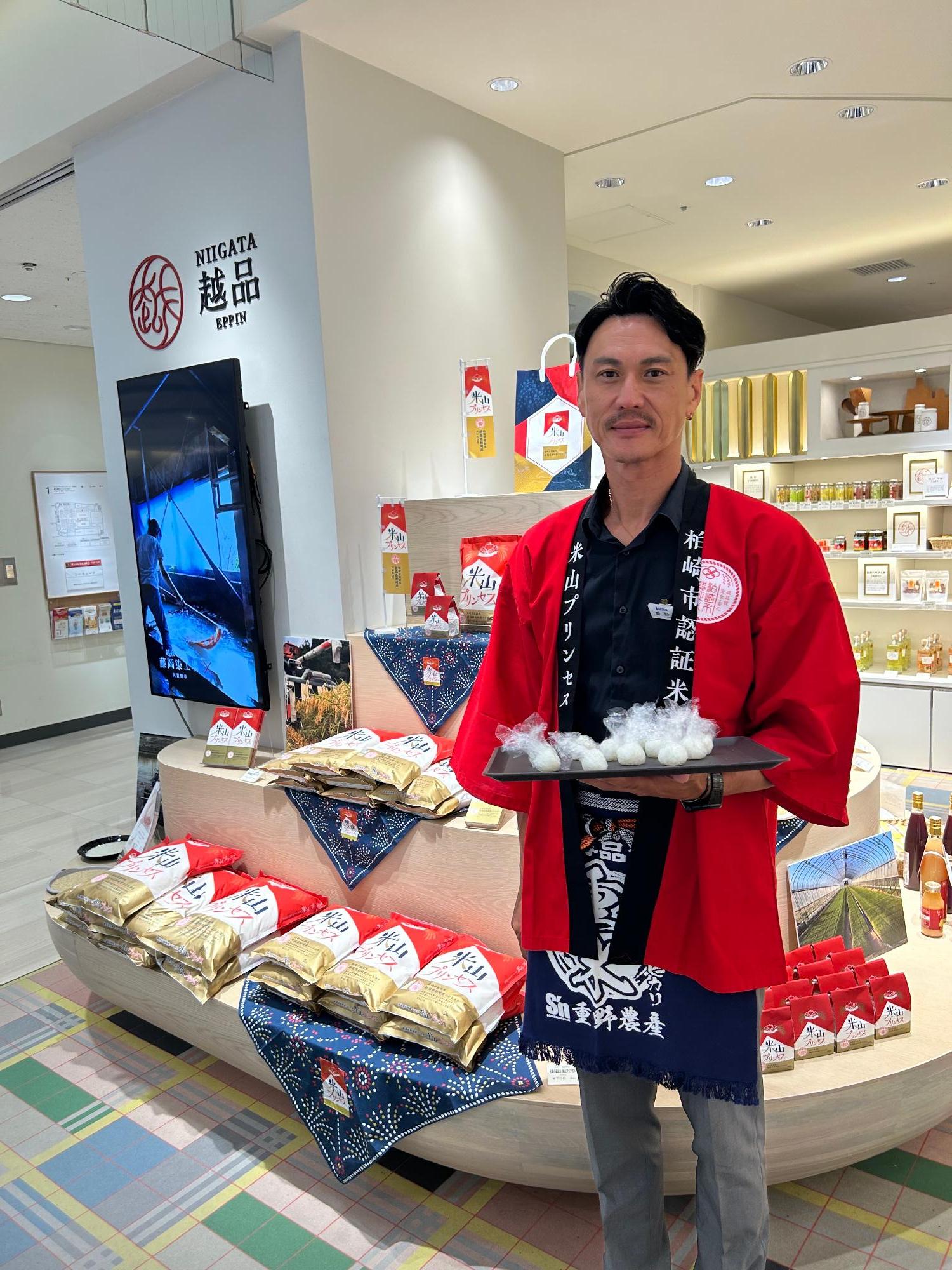 写真:新宿伊勢丹で、一口サイズに握られた米山プリンセスのおにぎりを配布する生産者の重野さん。重野さんは米山プリンセスと書かれた赤いハッピを着ています
