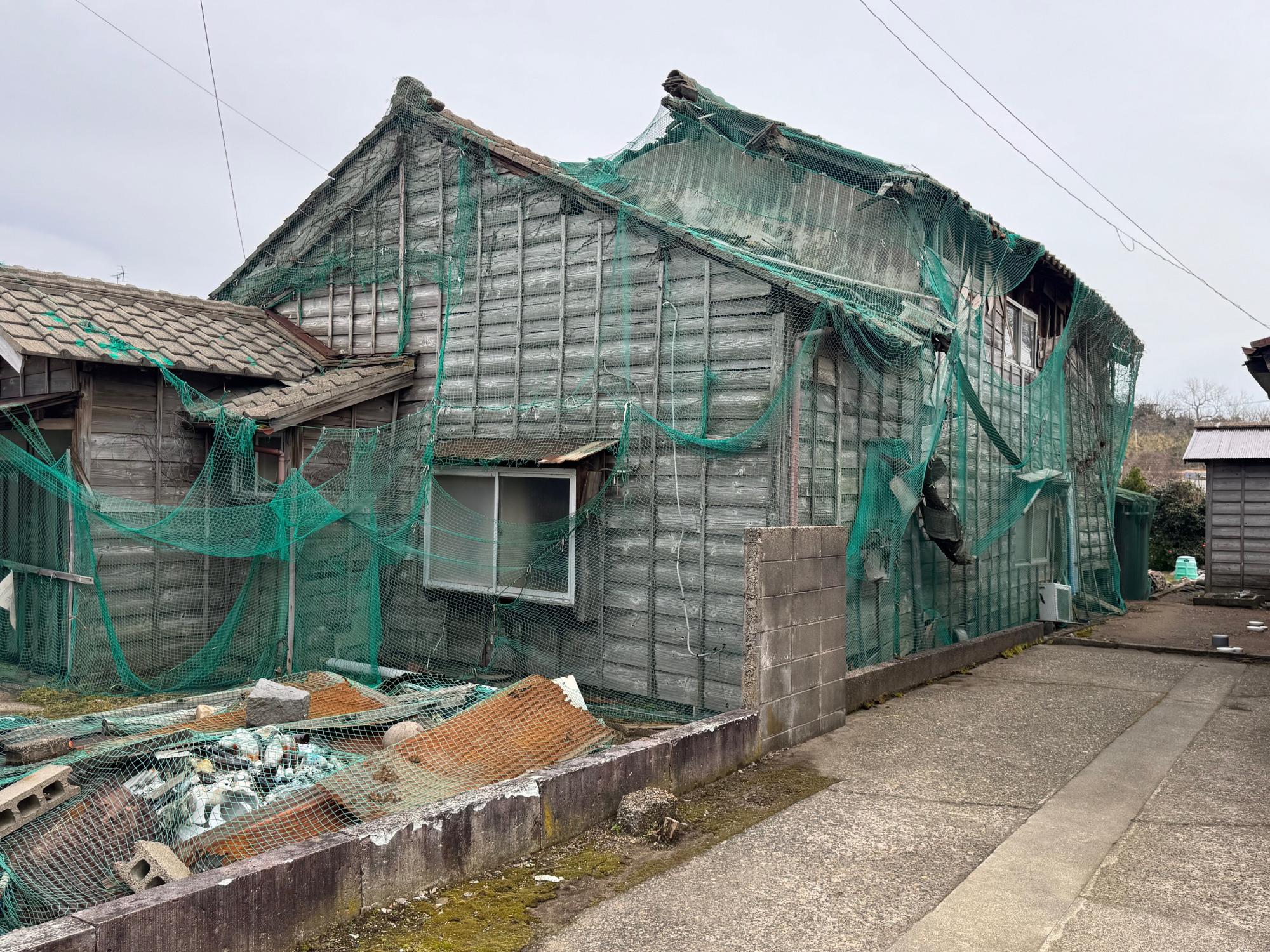 柏崎市荒浜三丁目地内特定空家の写真。2階部の軒部が損壊し、飛散防止用のネットが建物全体を覆っています