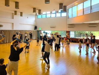 写真:参加者がパーティー体操用のタオルを両手首にかけななめに体を傾けています