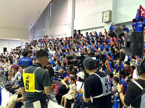 写真:11年ぶりに柏崎市で開催された日本選手権の様子を撮影するスタッフ。両手にバルーン型応援グッズを持ち、ブルボンKZを応援する観客席にカメラを向けています