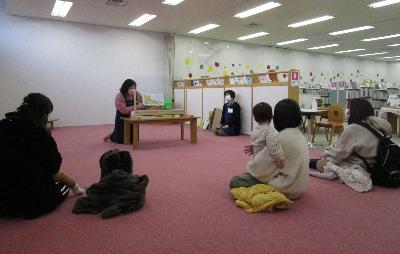 お母さんに抱っこされた小さい子どもたちが距離をとって座り、係の方が読んでくれる絵本を見ている様子の写真
