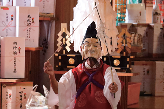 写真:笑みをたたえた男性のお面を身に着け、竹の釣竿をもって舞う様子