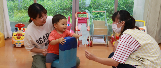 写真:椅子のおもちゃに座った子どもが子育て支援室の先生と遊んでいるところ。子どもの後ろにはお母さんが寄り添っています。