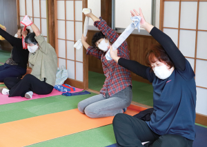 写真:タオルの両端を持った手を頭の上に上げ、体の脇を伸ばす体操をする参加者