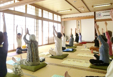 写真：座布団の上にあぐらをかき、先生の動きに合わせて、手のひらを頭の上で合わせて上に伸ばすポーズをしています