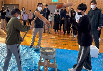 写真：大人が見守る中、2人の子どもが杵と臼でお餅をついています