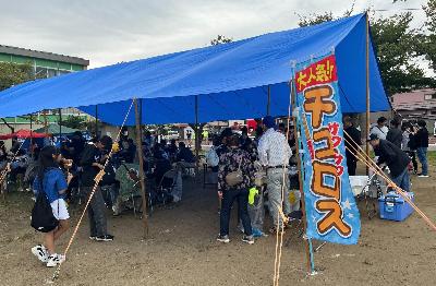 写真：コミセン隣の東部公園に設置されたブルーシート屋根の飲食スペース。屋根の下でたくさんの地域住民が、集まっています
