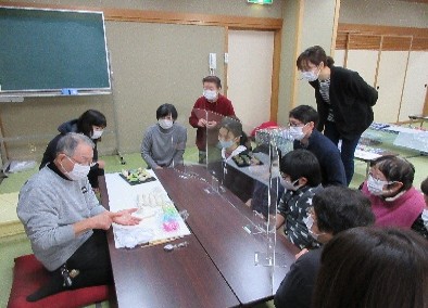 写真：先生の手の上で形作られていく「いんころ」を、じっと見つめる10人ほどの参加者。