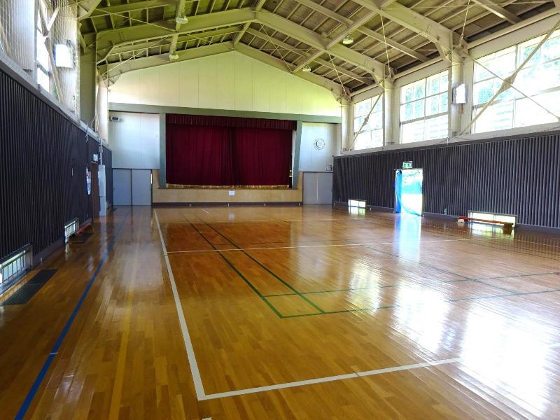 写真：講堂（体育館）。前方にステージがあり、バレーボールなどのラインテープが貼ってあります