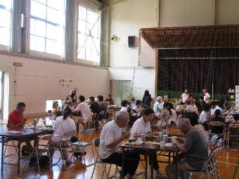 写真：コミセンの体育館に机といすが並べられ、大勢の人が会食しています