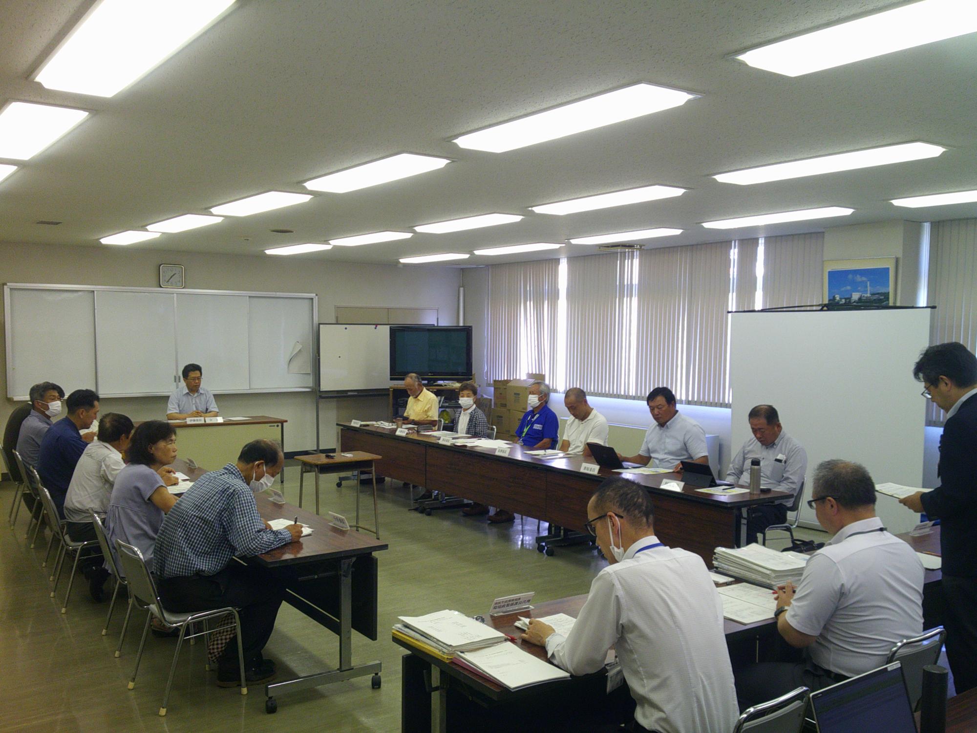 写真：会議室にロの字型に机を配置し、委員らが向かい合って報告を聞いています。