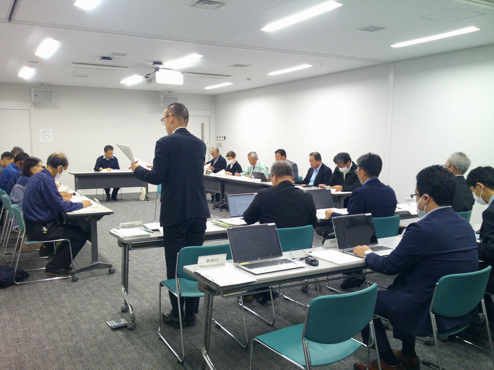 写真：会議室にロの字型に机を配置し、委員らが向かい合って報告を聞く様子。