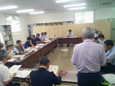 写真：会議室にロの字型に机を配置し、委員らが向かい合って報告を聞いています。1人の男性が起立し、報告しています。