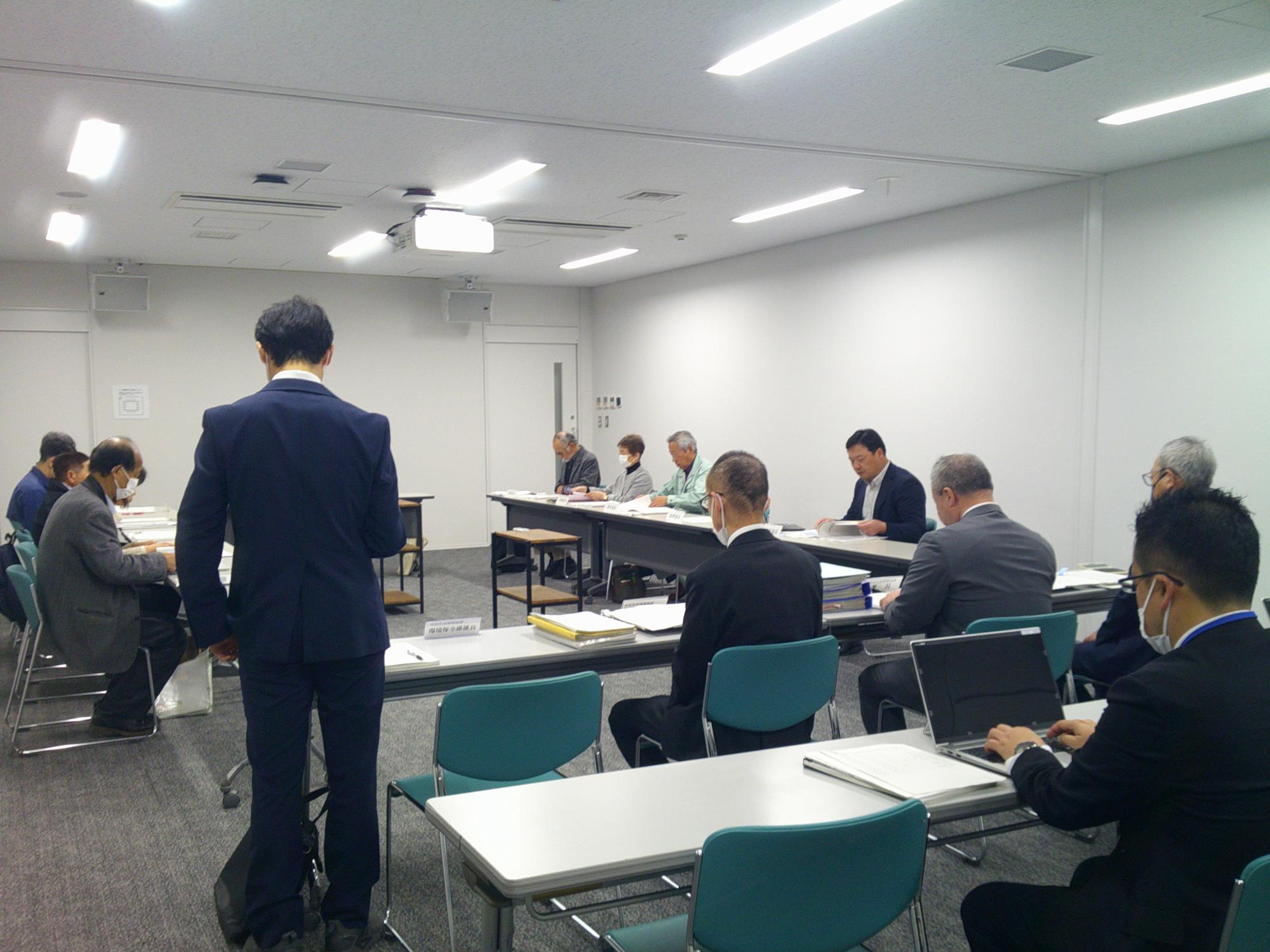 写真：会議室にコの字型に机を配置し、委員らが向かい合って報告を聞く様子。