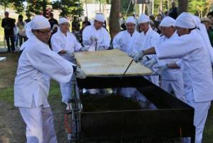 7人の白装束の男の人が四角い油の入った鍋に大きな油揚げの生地を入れようとしているところの写真