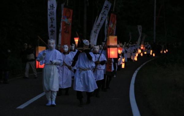 夕暮れの道を、白い衣装とお面で狐に扮した人たちが赤や白ののぼりとちょうちんを持ち2列の行列をつくって歩いている写真