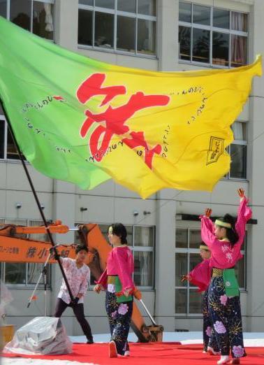 舞と書かれた大きな旗を中学生の男子が振る前で、濃い桃色が入った上着に緑色の衣装を着た女性がよさこいを舞っている写真