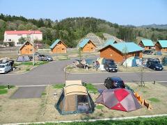 青空の下に水色の屋根のログキャビンが並び、手前には芝生のオートキャンプサイトに複数のテントが並んでいる写真