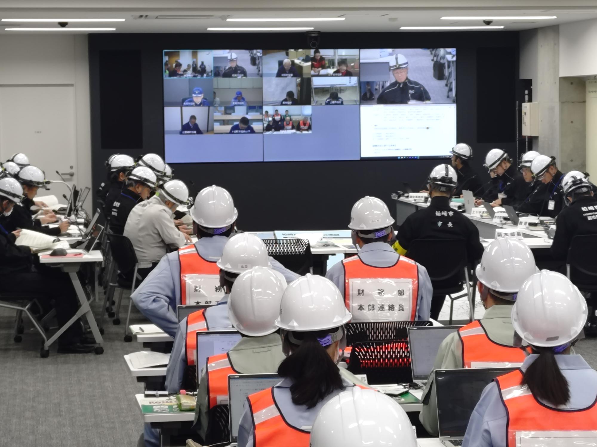 写真：テレビ会議で県や他市町村と協議を行っている様子。前面のモニターには別自治体の対策本部の映像が写っています