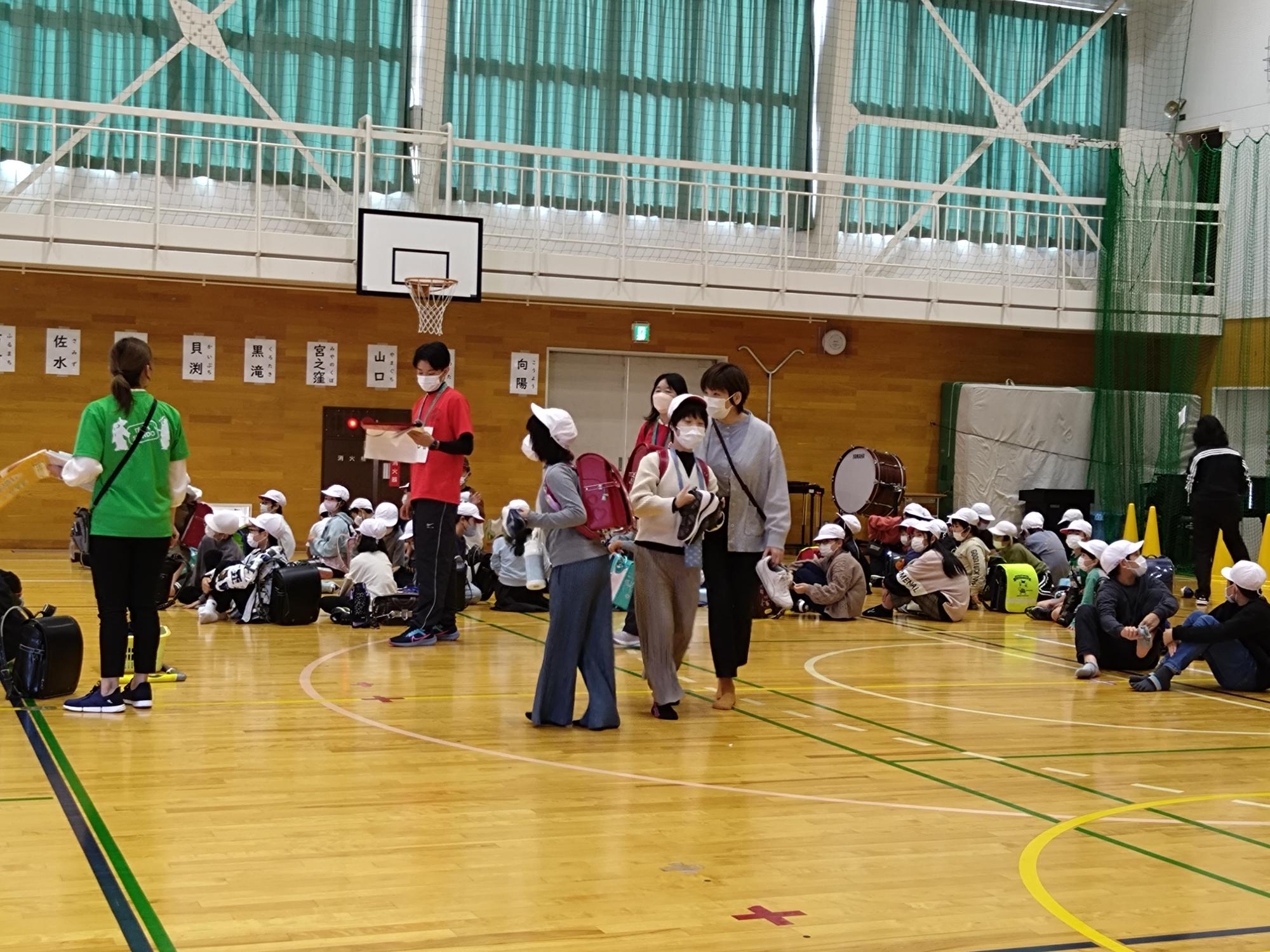 写真：学校の体育館で並んで保護者の迎えを待つ児童。教職員らが引き渡し業務にあたっています