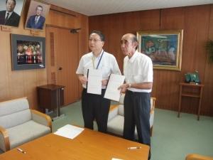 協定書を両手に持って、机の前に並んで立つ南日会長と会田市長の写真