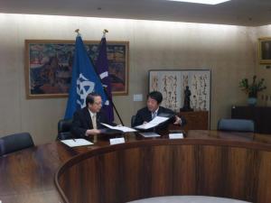 互い違いに掲げられる2本の旗前の円形の机に座り、協定書を取り交わす会田市長と山本前橋市長の様子の写真