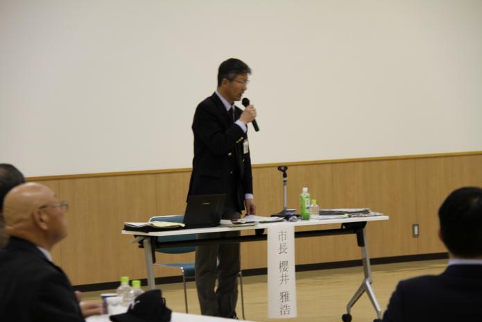 意見交換会会場の参加者の方々の前に設置された市長用の机の前で右手にマイクを持ち質問に答える櫻井市長のアップの写真