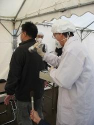 テント内で白衣にマスクをつけた職員が後ろ向きの男性の背中に機械を当てスクリーニングを実施している様子の写真