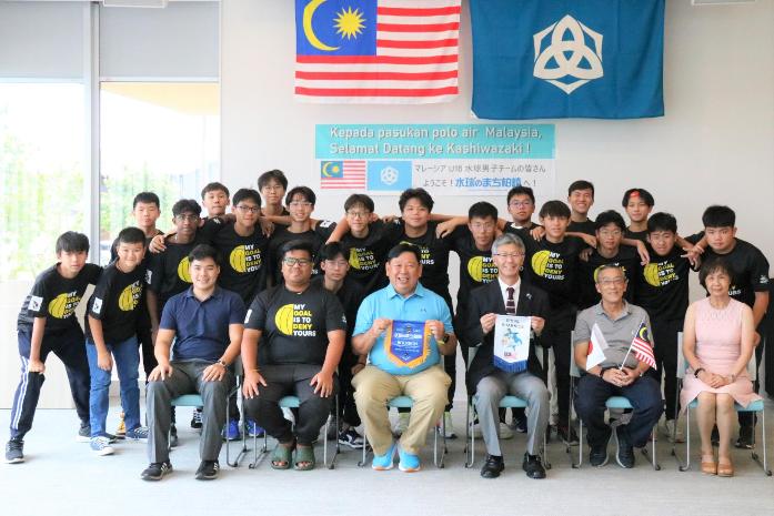 市長がペナントを持って、マレーシアの水球男子ジュニアチームと並んで記念撮影している様子