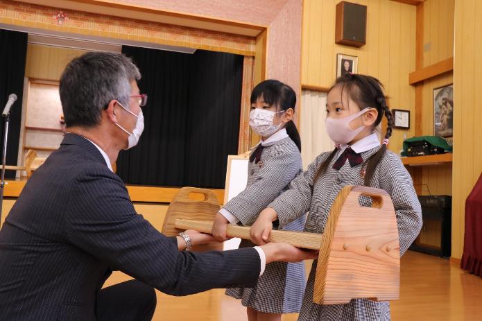 柏崎産の杉で作った木製ベンチを市長が幼稚園児2人に両手で手渡している様子