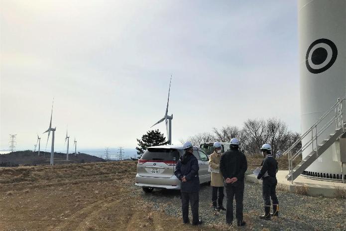 風力発電用の風車が立ち並ぶ敷地で、ヘルメットを被った市長が説明を受けているところ