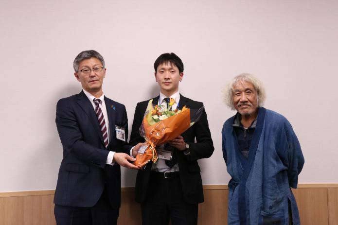市長が地域おこし協力隊の小柴さんに花束を手渡し、世話人である小林さんと3人で並び記念撮影をしている