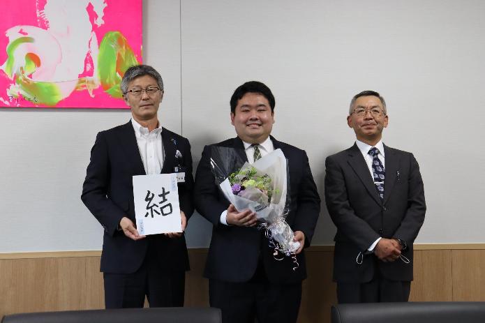 市長が地域おこし協力隊の坪井さんに花束を渡し、世話人の池田さんと3人で並び記念撮影をしている