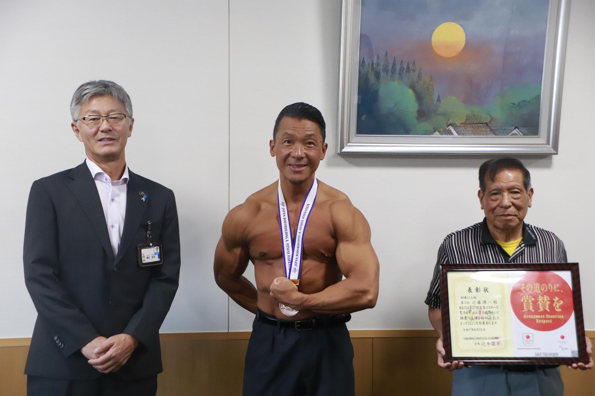 写真：市長（左）と首からメダルを下げた近藤さん（中央）。近藤さんは鍛えた筋肉美が見えるように、上半身裸です。近藤さんの右側にはトレーニング仲間の梅沢さんが賞状を持っています