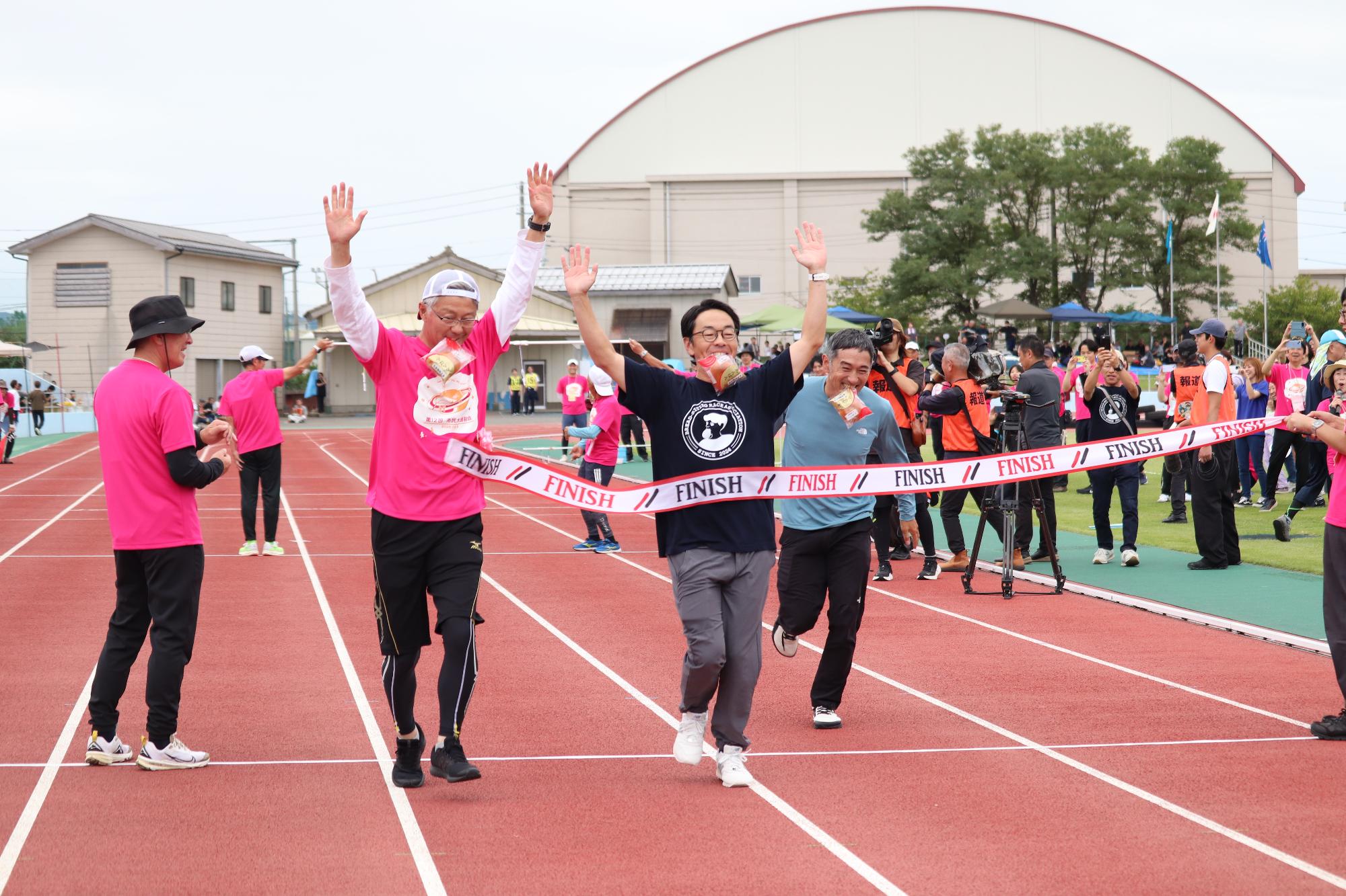 写真：パン食い競争に参加した市長。パンを口にくわえたまま、ピンク色のTシャツを着た市長がゴールテープを切っています