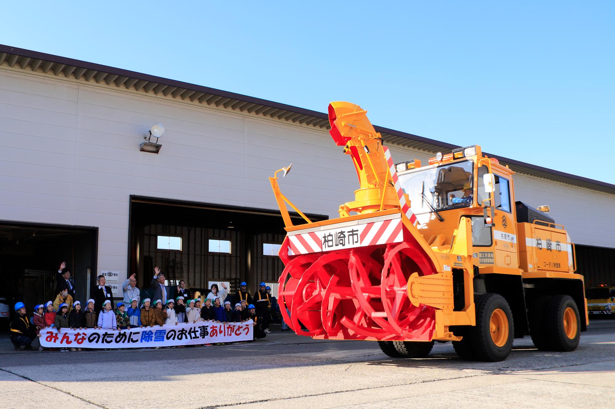 写真：除雪車の出動式に出席した市長。子どもたちと一緒に除雪車に向かって手を振っています。