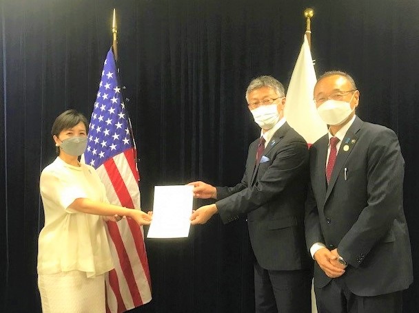 日本とアメリカの両国旗の前で、桜井柏崎市長と佐渡市長が米国大使館書記官へ要望書を手渡している写真