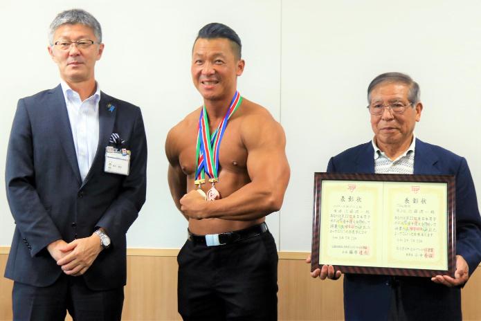 メダルを肩にかけ鍛え上げた筋肉を披露する近藤さんと賞状を持った同席者の梅沢さん、櫻井柏崎市長が横に並んでいる写真