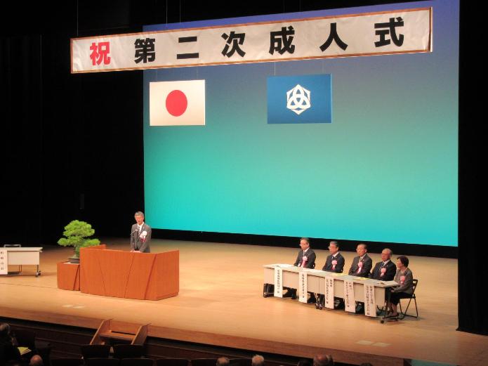 第二次成人式と書かれた横看板の下に国旗と市旗が掲げられたステージ上であいさつをする櫻井市長