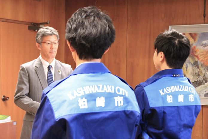 青い制服を着た2人の男性職員が市長の前に立ち市長から声をかけられている様子