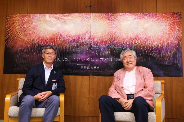 片貝煙火工業の本田社長と市長が花火のポスターの前で並んで椅子に座っている
