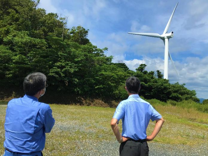 自然豊かで広大な土地にそびえ立つ風力発電施設を見上げている市長の後ろ姿の写真
